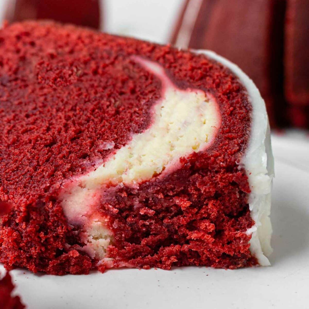 A close-up of a slice of red velvet pound cake with a creamy white cheesecake filling and white icing on the outside, set on a white plate.