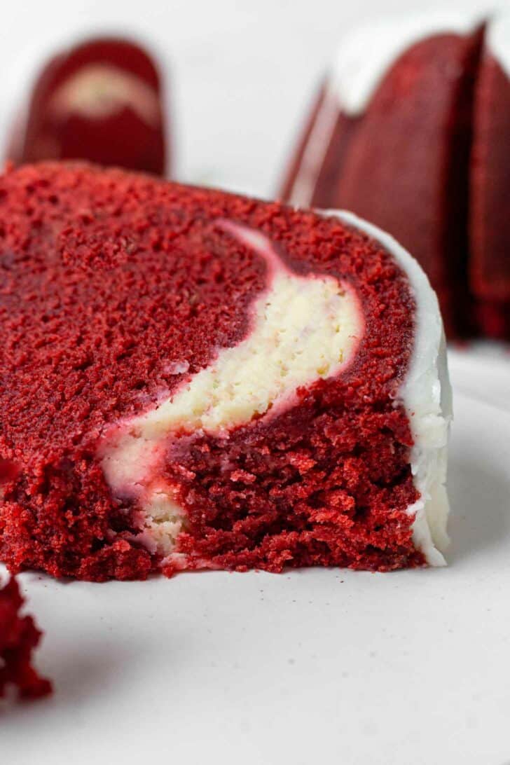 A close-up of a slice of red velvet pound cake with a creamy white cheesecake filling and white icing on the outside, set on a white plate.