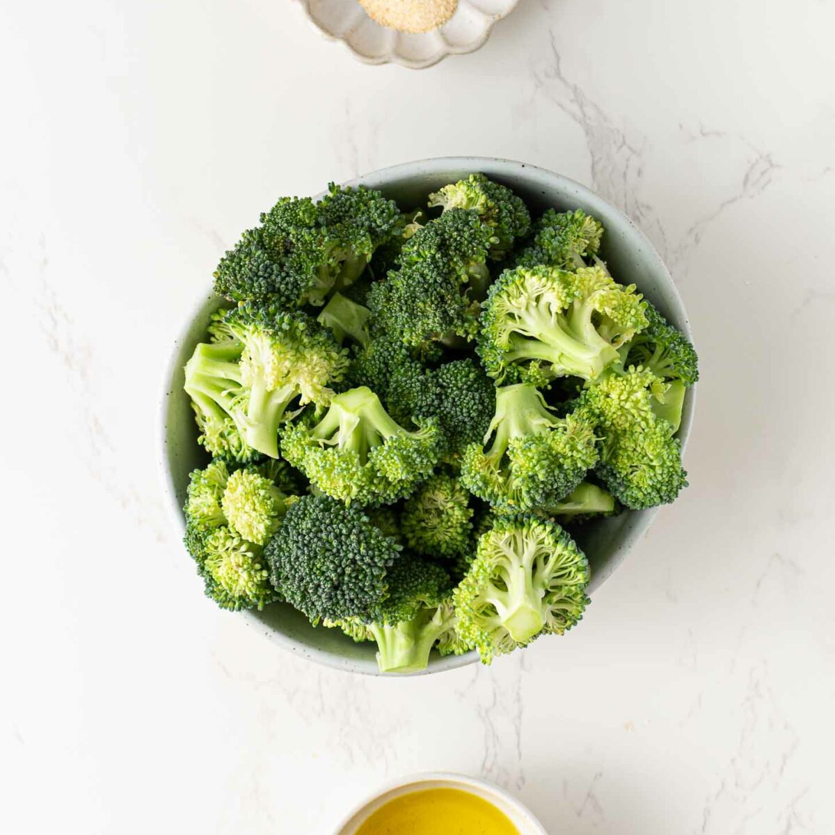 A bowl of fresh broccoli florets, a small dish of olive oil, and a scalloped dish containing salt, pepper, and granulated seasonings are arranged on a white marble surface.