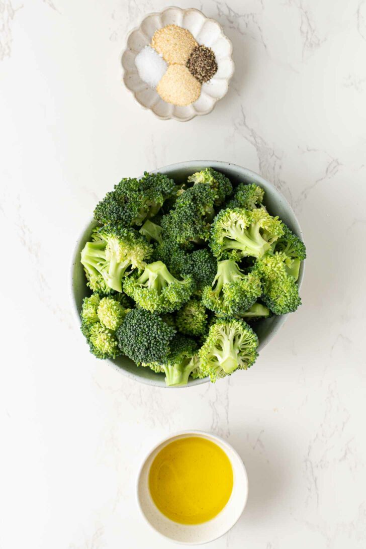 A bowl of fresh broccoli florets, a small dish of olive oil, and a scalloped dish containing salt, pepper, and granulated seasonings are arranged on a white marble surface.