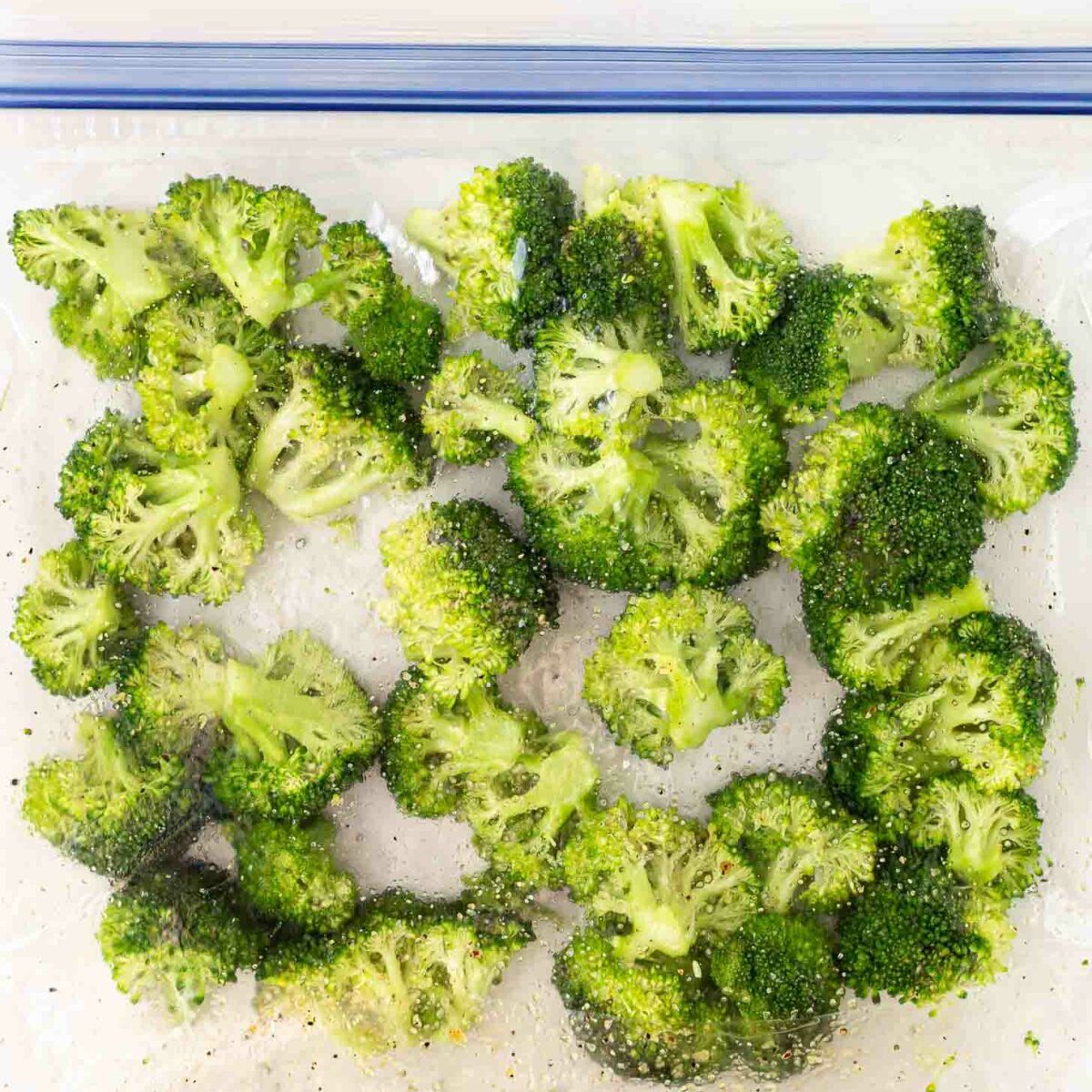 Broccoli florets coated with seasoning inside a large clear resealable plastic bag, viewed from above on a white background.