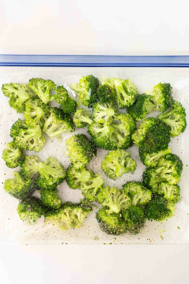 Broccoli florets coated with seasoning inside a large clear resealable plastic bag, viewed from above on a white background.