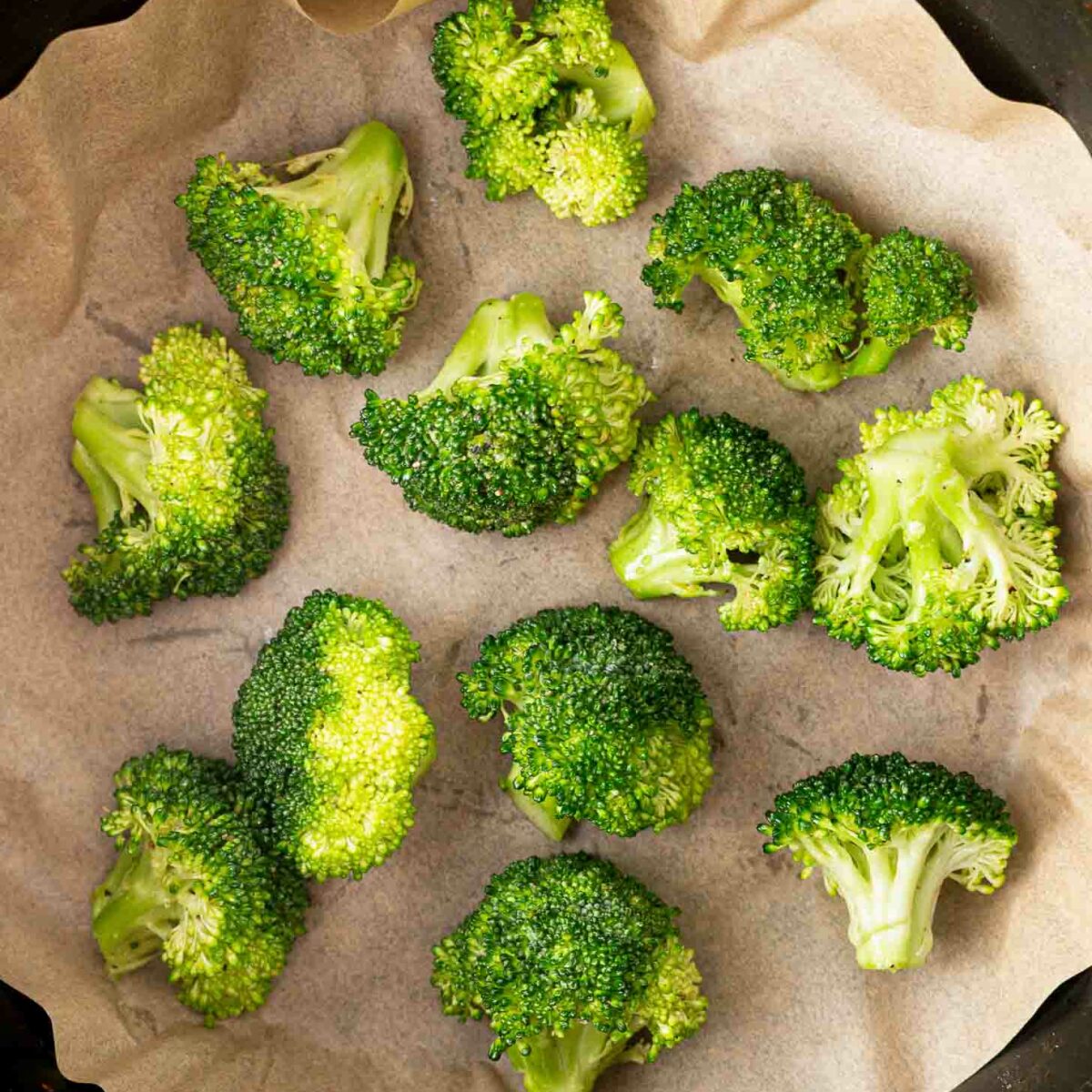 Fresh broccoli florets arranged on a sheet of parchment paper inside a black round air fryer basket, ready to be cooked.