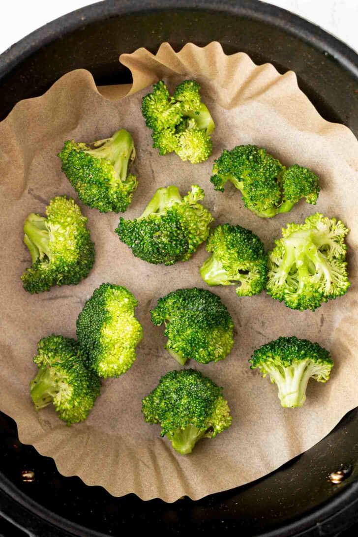 Fresh broccoli florets arranged on a sheet of parchment paper inside a black round air fryer basket, ready to be cooked.
