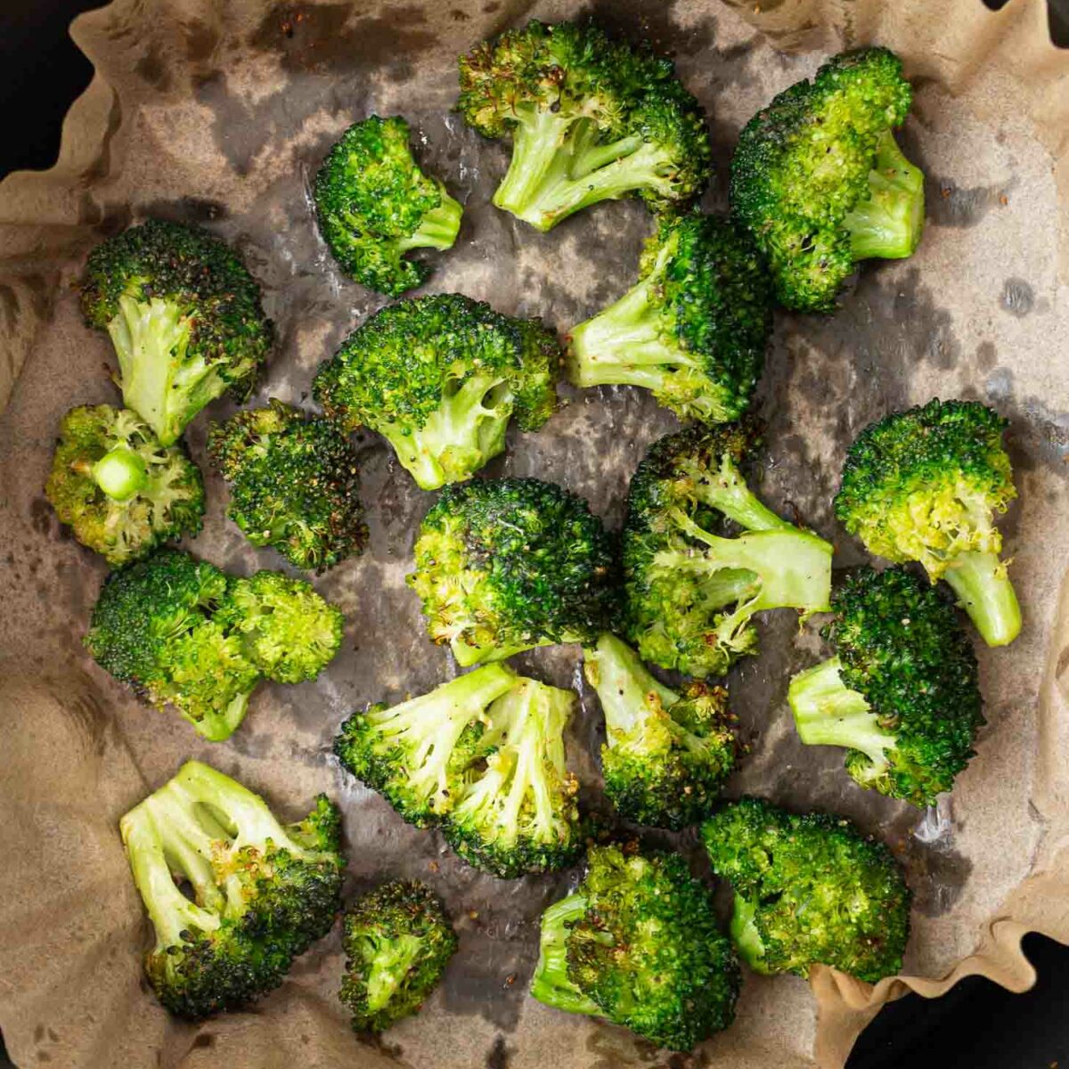 Broccoli florets cooked and slightly browned on parchment paper inside a black air fryer basket.