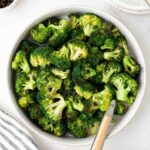 A bowl filled with roasted broccoli florets sits on a white surface beside a fork, a striped towel, and small bowls of ground pepper and breadcrumbs.