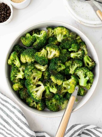 A bowl filled with roasted broccoli florets sits on a white surface beside a fork, a striped towel, and small bowls of ground pepper and breadcrumbs.