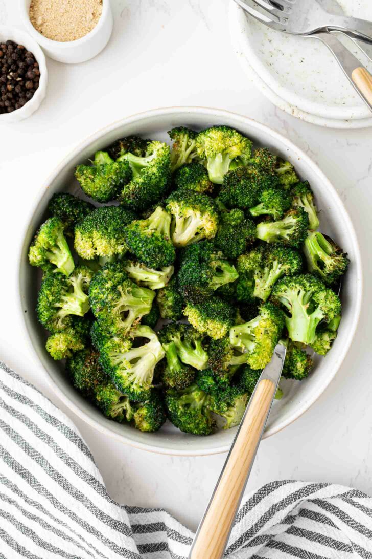A bowl filled with roasted broccoli florets sits on a white surface beside a fork, a striped towel, and small bowls of ground pepper and breadcrumbs.