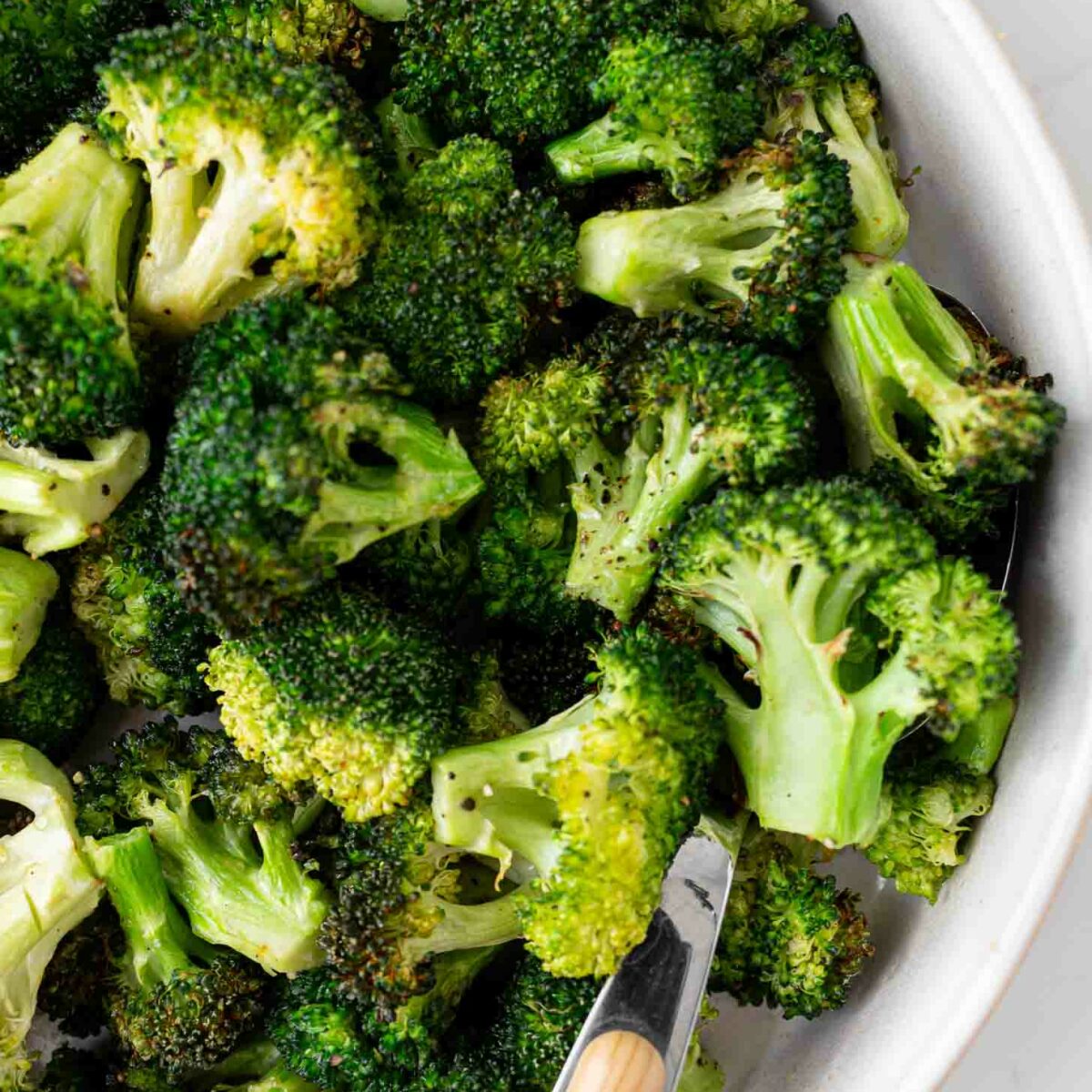 A close-up of roasted broccoli florets in a white bowl with a wooden-handled serving utensil. The broccoli is slightly browned, indicating it has been roasted.