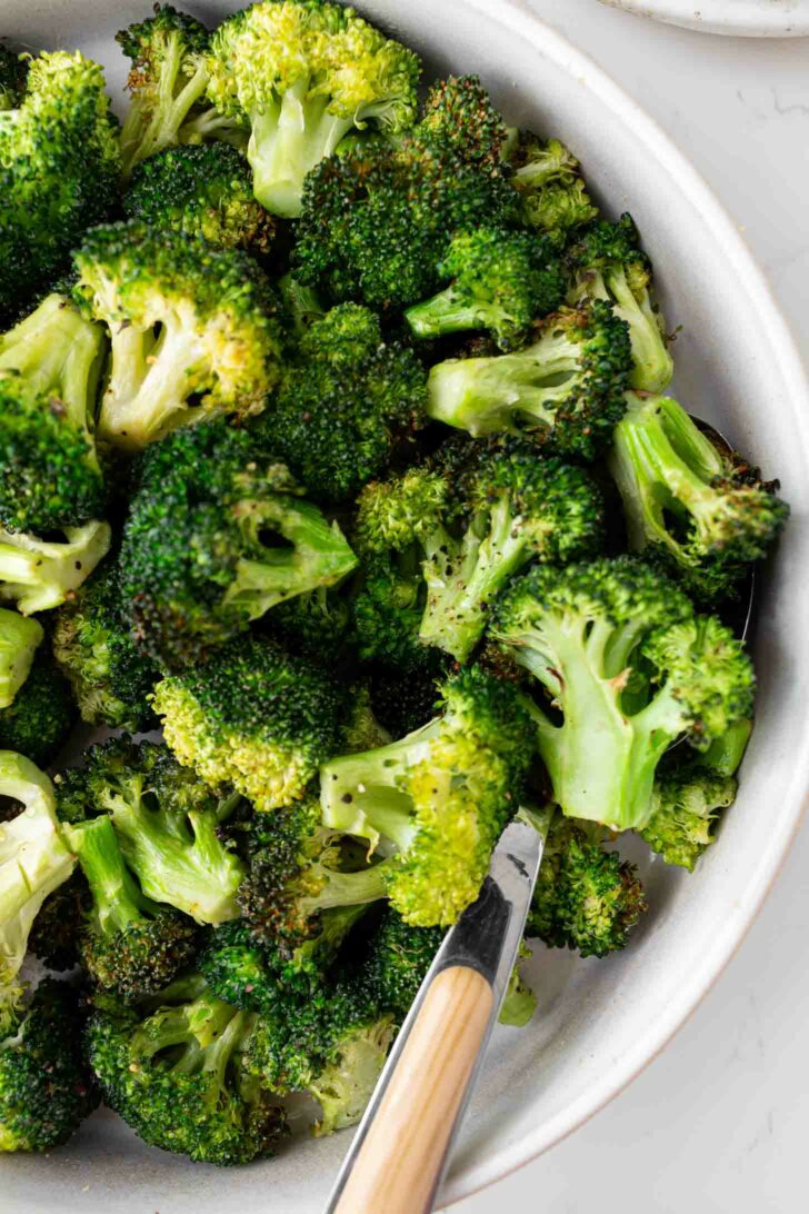 A close-up of roasted broccoli florets in a white bowl with a wooden-handled serving utensil. The broccoli is slightly browned, indicating it has been roasted.