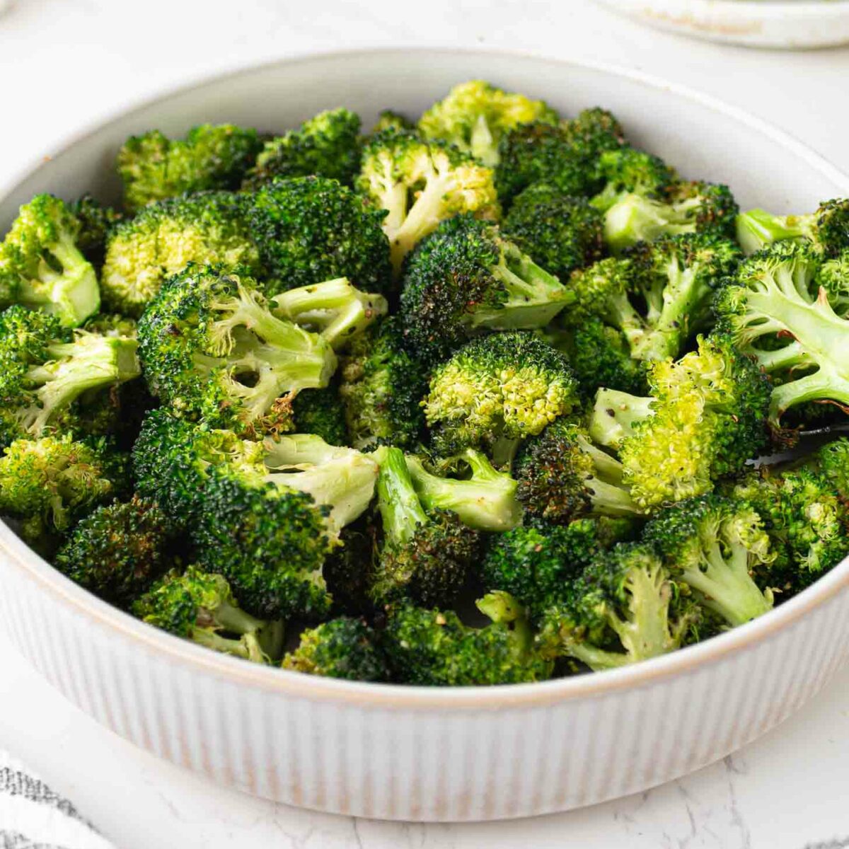 A bowl filled with roasted broccoli florets sits on a white marble surface, with a stack of plates and forks in the background and a striped cloth nearby.