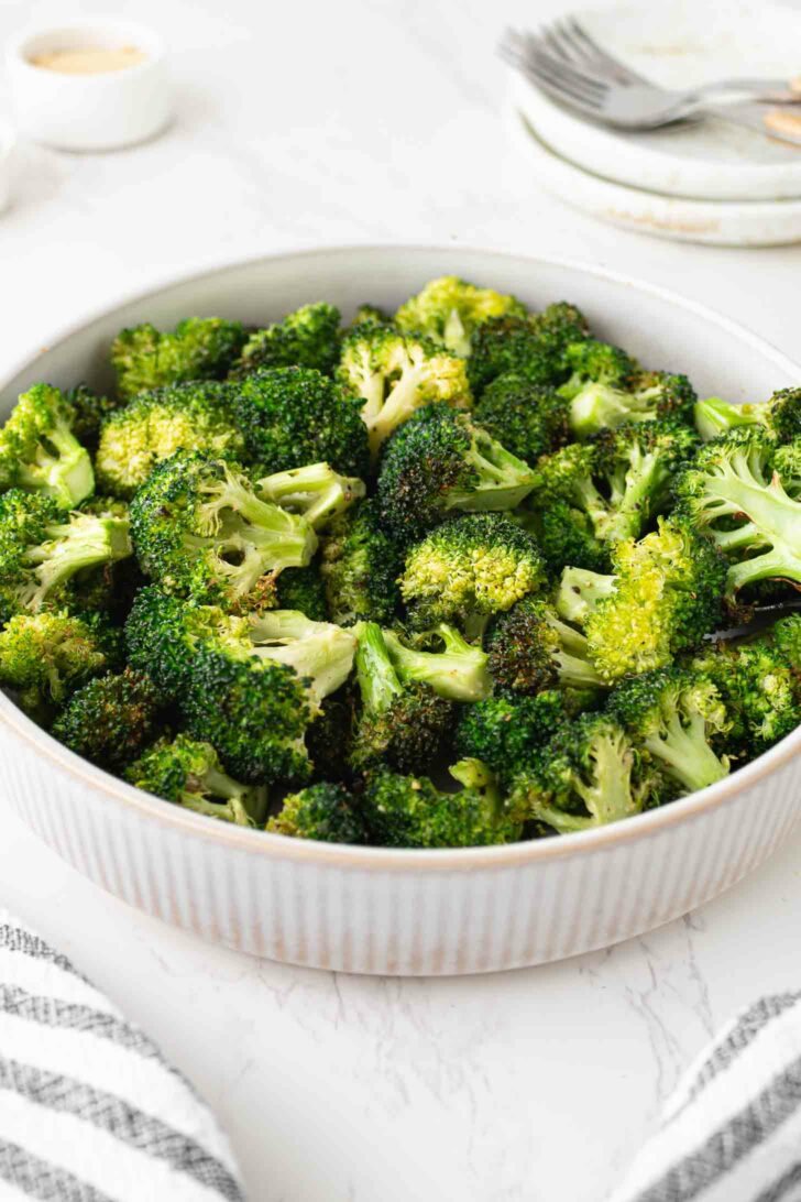 A bowl filled with roasted broccoli florets sits on a white marble surface, with a stack of plates and forks in the background and a striped cloth nearby.