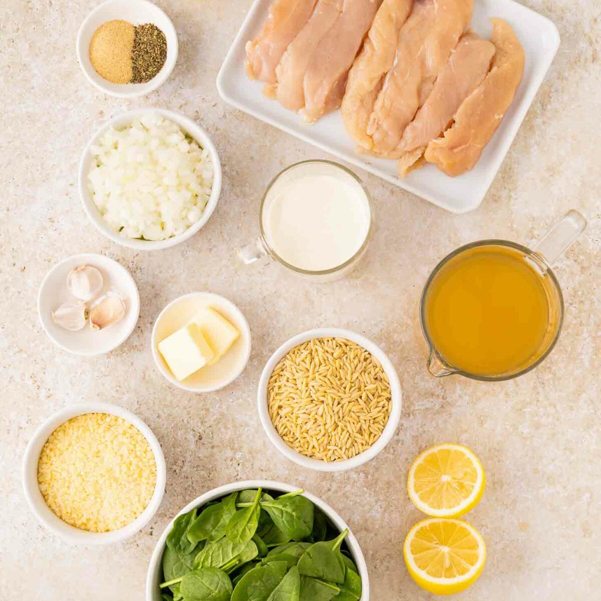 Overhead view of raw chicken breasts, olive oil, chopped onions, garlic cloves, spices, butter, cream, chicken broth, uncooked orzo, grated Parmesan, fresh spinach, and halved lemons on a light countertop.