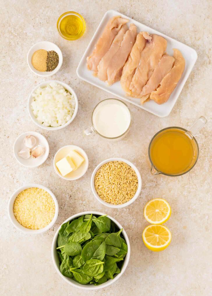 Overhead view of raw chicken breasts, olive oil, chopped onions, garlic cloves, spices, butter, cream, chicken broth, uncooked orzo, grated Parmesan, fresh spinach, and halved lemons on a light countertop.