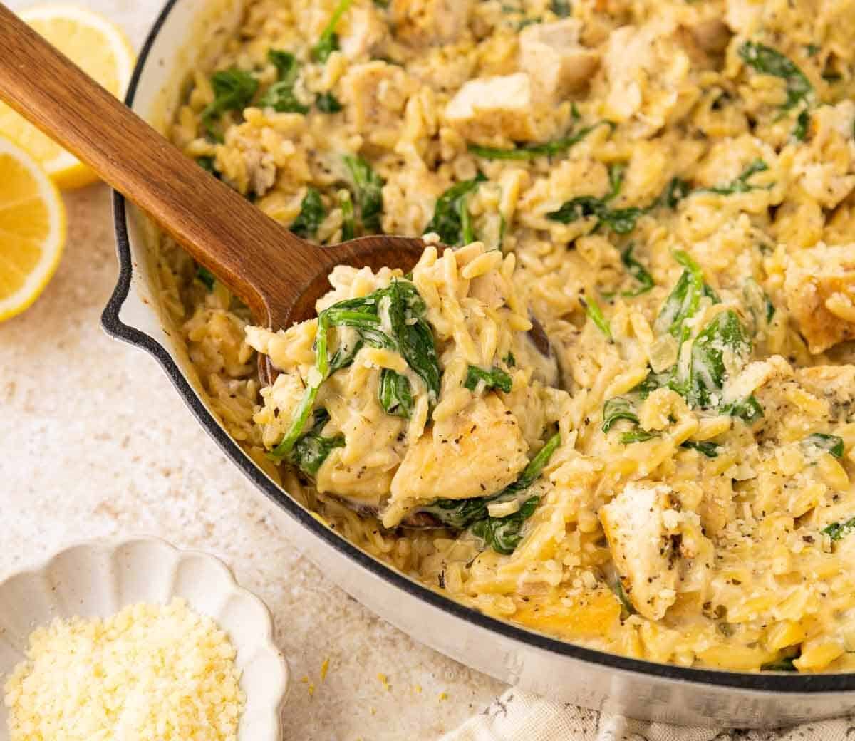 A skillet filled with creamy orzo pasta, chunks of chicken, spinach, and grated cheese; a wooden spoon scoops some up. Sliced lemons and a small bowl of grated cheese are nearby.