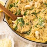 A skillet filled with creamy orzo pasta, chunks of chicken, spinach, and grated cheese; a wooden spoon scoops some up. Sliced lemons and a small bowl of grated cheese are nearby.