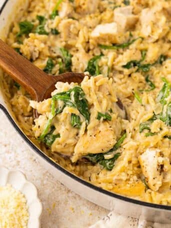 A skillet filled with creamy orzo pasta, chunks of chicken, spinach, and grated cheese; a wooden spoon scoops some up. Sliced lemons and a small bowl of grated cheese are nearby.