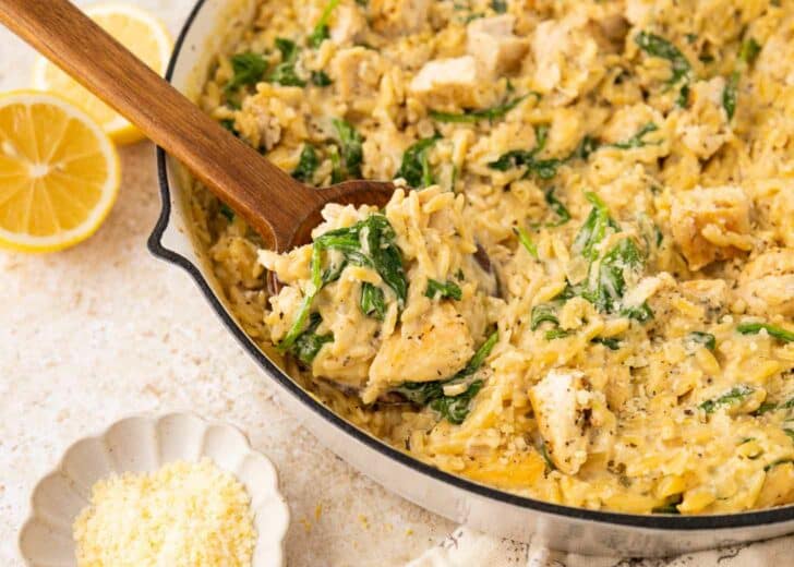 A skillet filled with creamy orzo pasta, chunks of chicken, spinach, and grated cheese; a wooden spoon scoops some up. Sliced lemons and a small bowl of grated cheese are nearby.