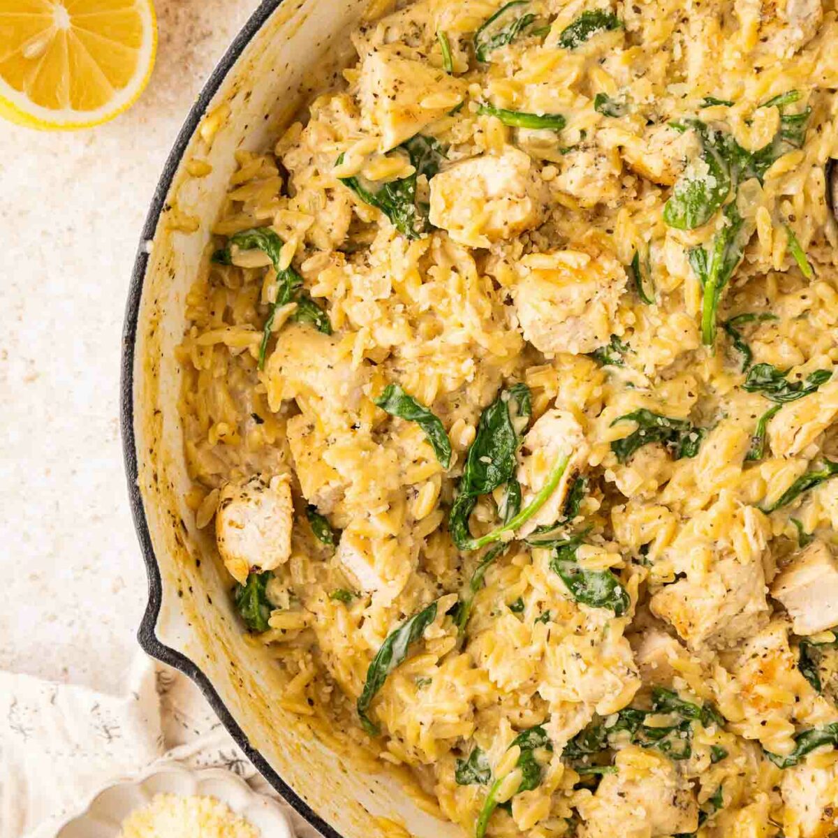A skillet filled with creamy orzo pasta, pieces of chicken, and spinach. The dish is garnished with black pepper. Next to the skillet are lemon halves and a small bowl of grated cheese.