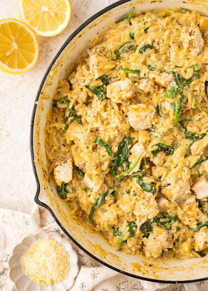 A skillet filled with creamy orzo pasta, pieces of chicken, and spinach. The dish is garnished with black pepper. Next to the skillet are lemon halves and a small bowl of grated cheese.