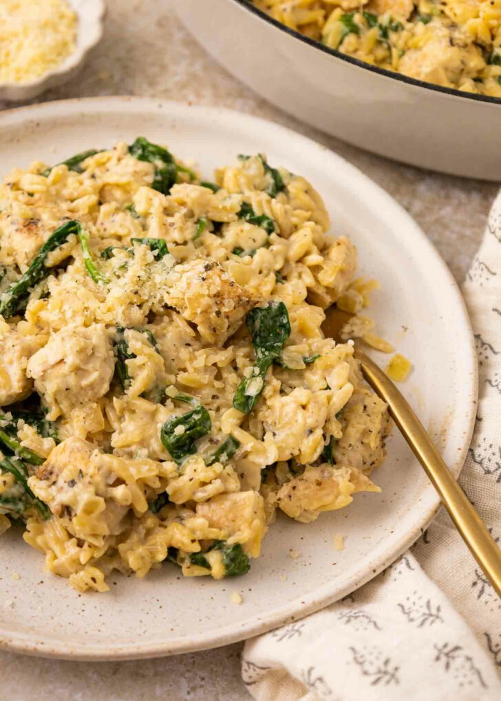 A plate of creamy chicken and spinach orzo garnished with grated cheese, served with a gold fork on a light-colored plate. A patterned napkin and a dish of orzo are visible beside the plate.
