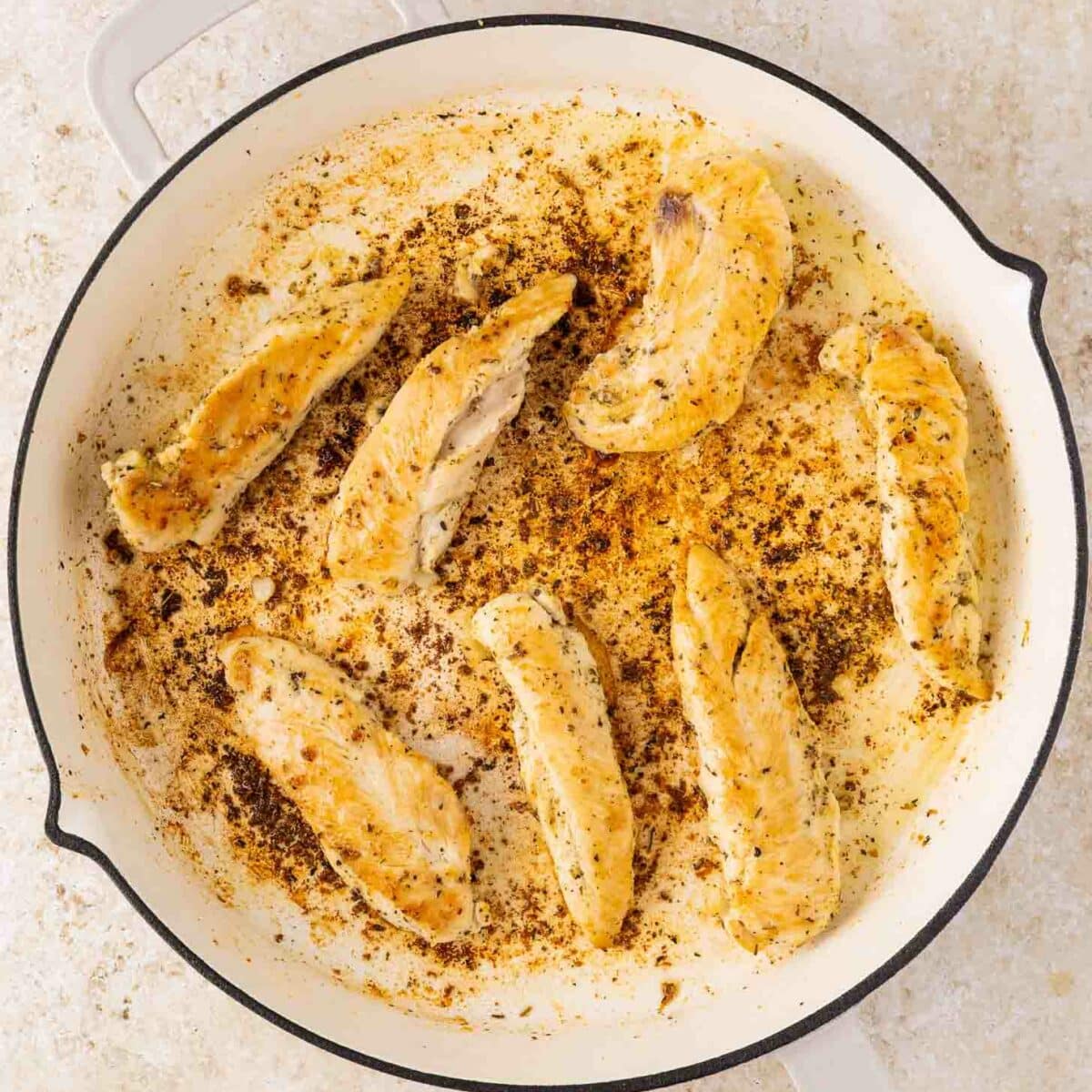 Seven cooked chicken tenders are arranged in a white skillet with browned seasoning and pan drippings visible. The skillet sits on a light-colored surface.