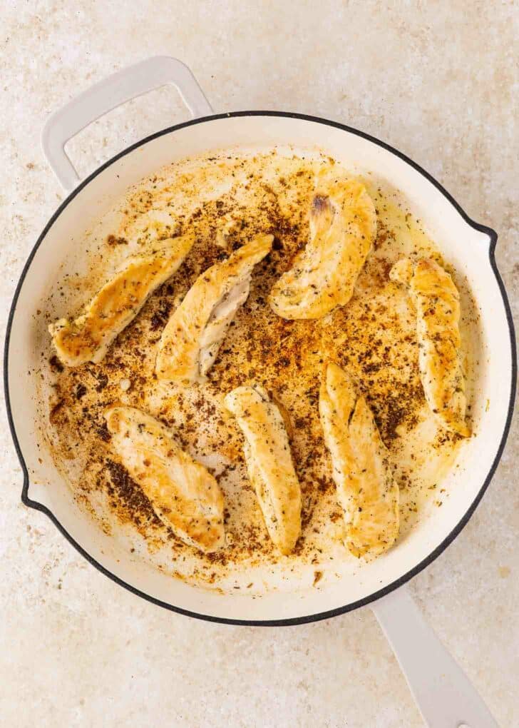 Seven cooked chicken tenders are arranged in a white skillet with browned seasoning and pan drippings visible. The skillet sits on a light-colored surface.