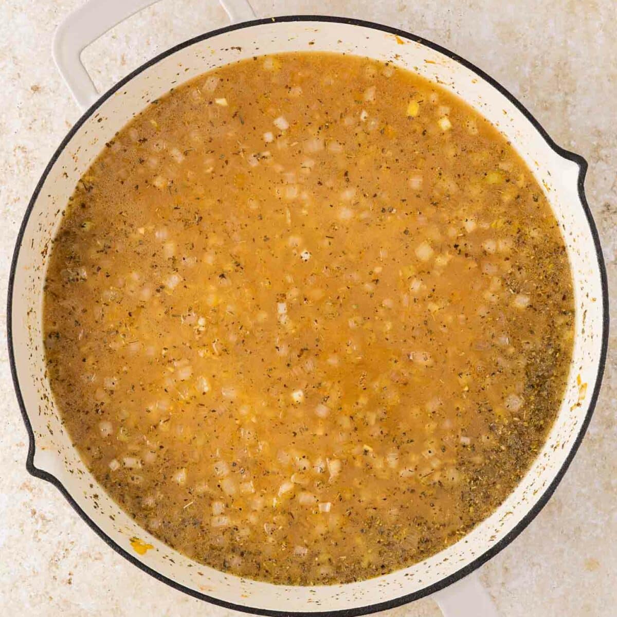 A large white pot filled with a light brown broth and orzo containing visible chopped onions and herbs, sitting on a light-colored countertop.
