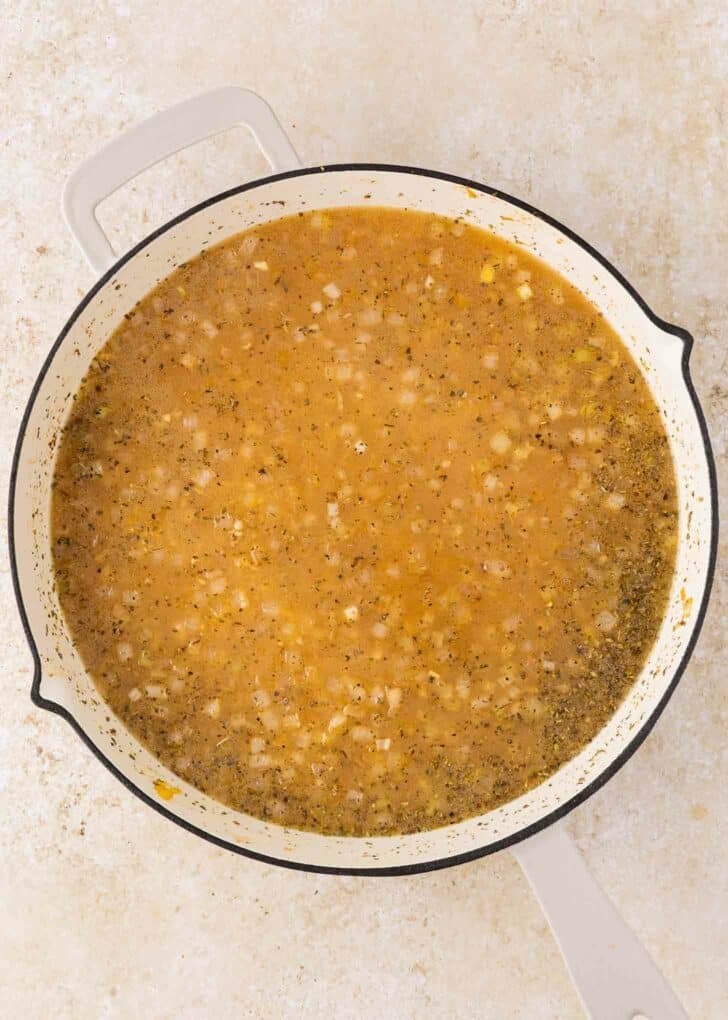 A large white pot filled with a light brown broth and orzo containing visible chopped onions and herbs, sitting on a light-colored countertop.