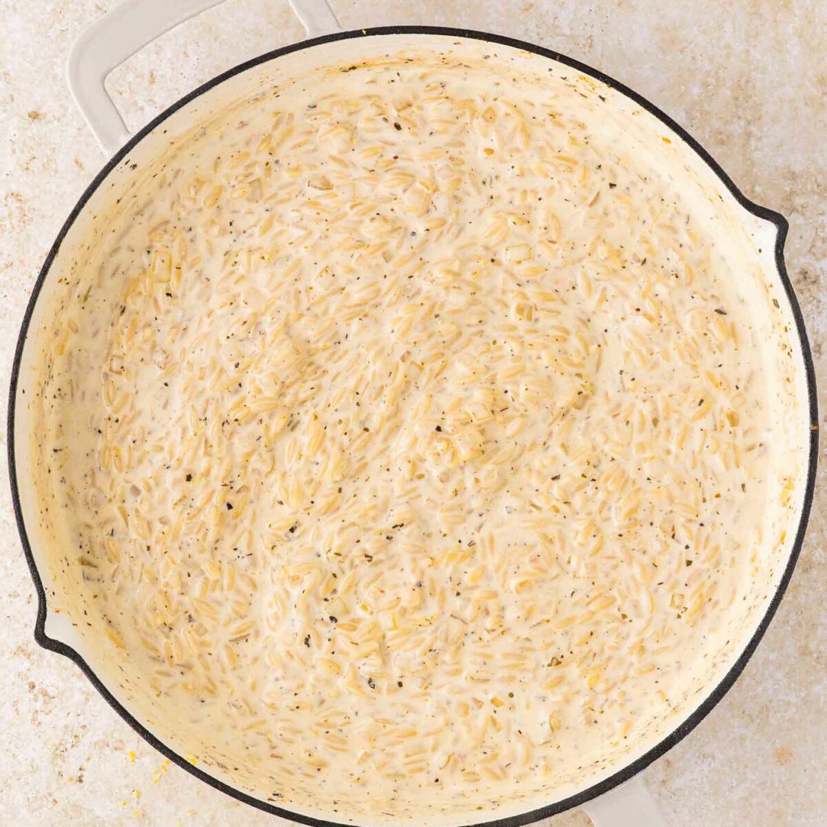 A large, shallow pan filled with creamy, cooked orzo pasta in a white sauce with visible herbs, sitting on a light-colored countertop.