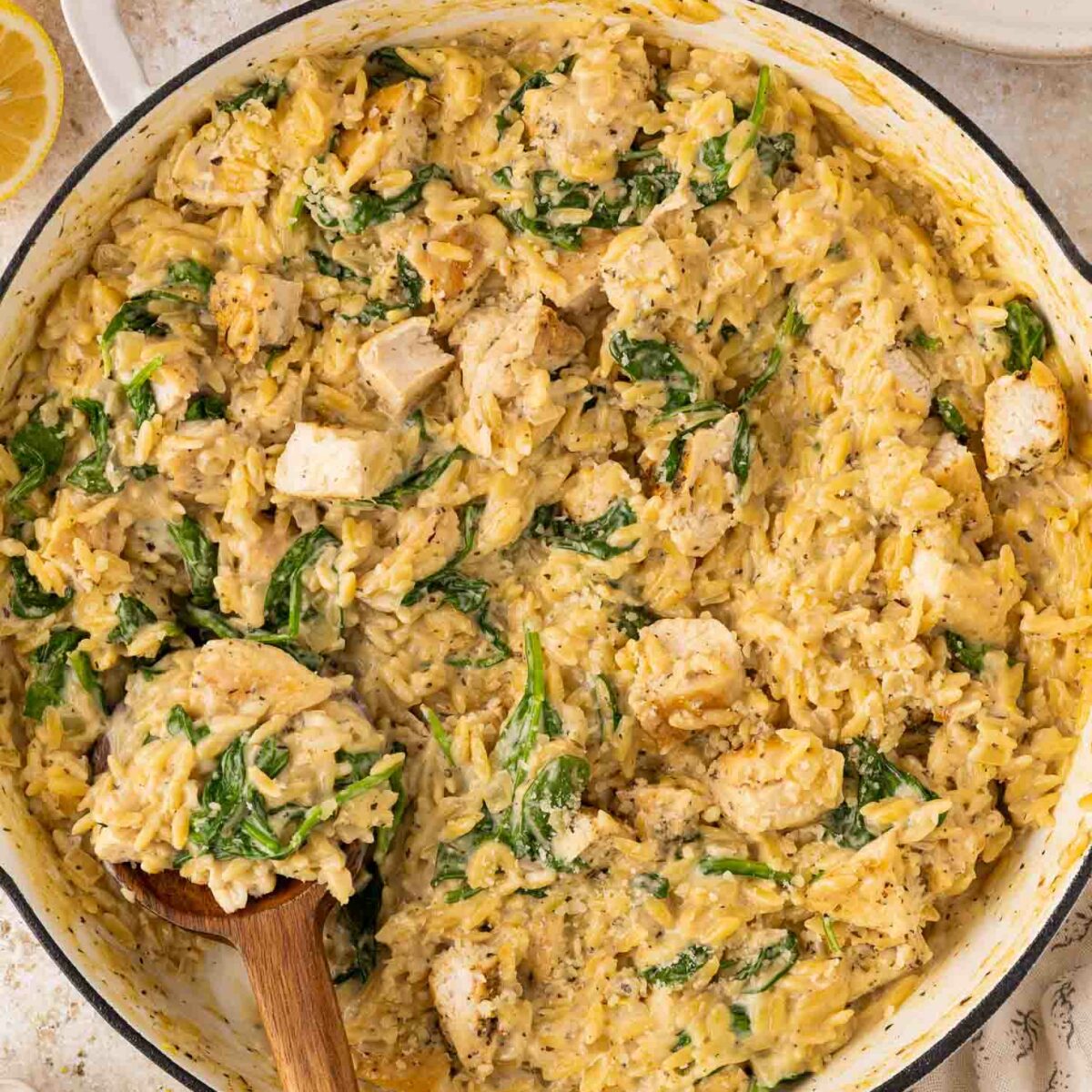 A large white skillet filled with creamy orzo pasta, pieces of cooked chicken, and fresh spinach. A wooden spoon is scooping up a portion. Slices of lemon and a plate are visible nearby.
