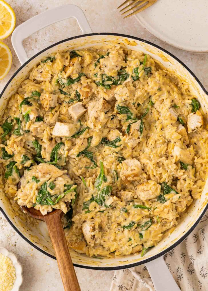 A large white skillet filled with creamy orzo pasta, pieces of cooked chicken, and fresh spinach. A wooden spoon is scooping up a portion. Slices of lemon and a plate are visible nearby.