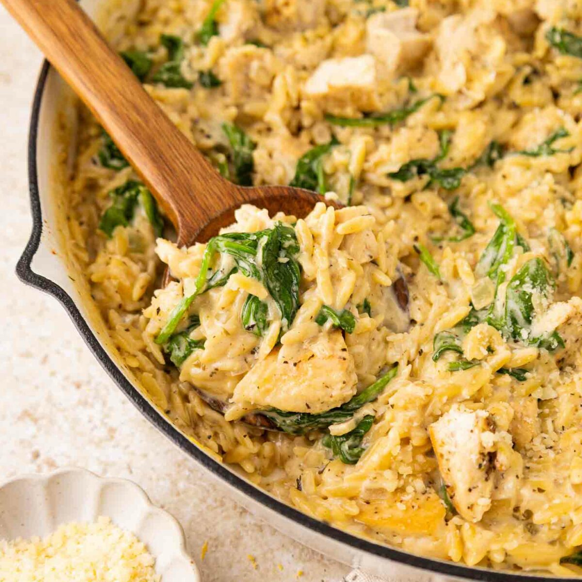 A creamy chicken and spinach orzo dish is being stirred with a wooden spoon in a skillet. Grated cheese is in a small bowl nearby, and a lemon wedge is visible in the background.