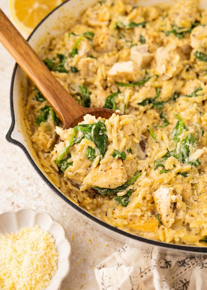 A creamy chicken and spinach orzo dish is being stirred with a wooden spoon in a skillet. Grated cheese is in a small bowl nearby, and a lemon wedge is visible in the background.
