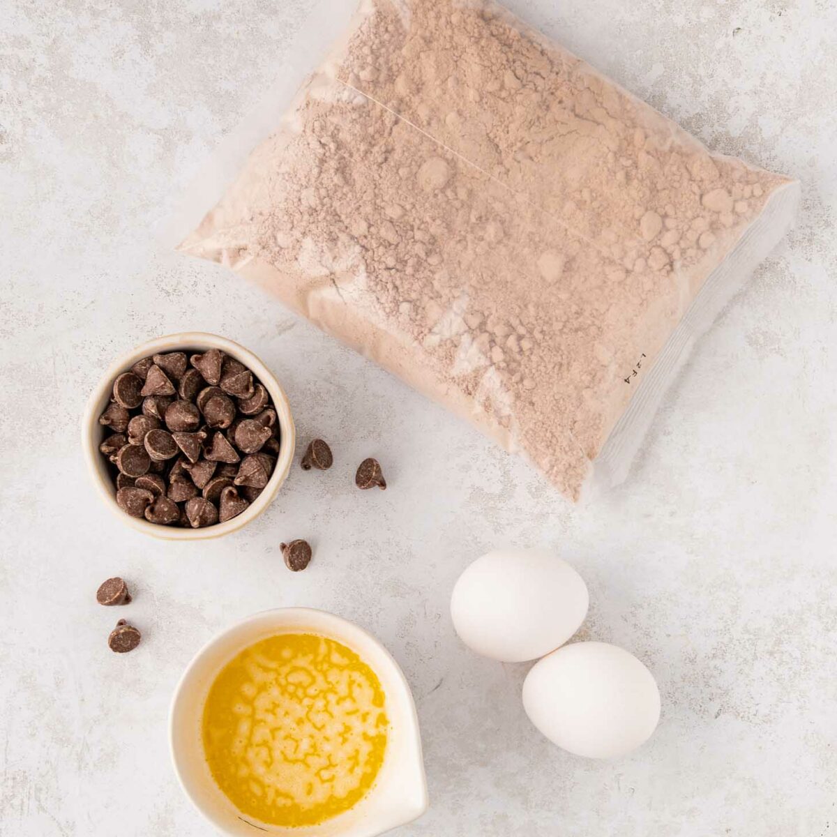 A bag of red velvet cake mix, a small bowl of chocolate chips, two eggs, and a small bowl of melted butter arranged on a light-colored surface.