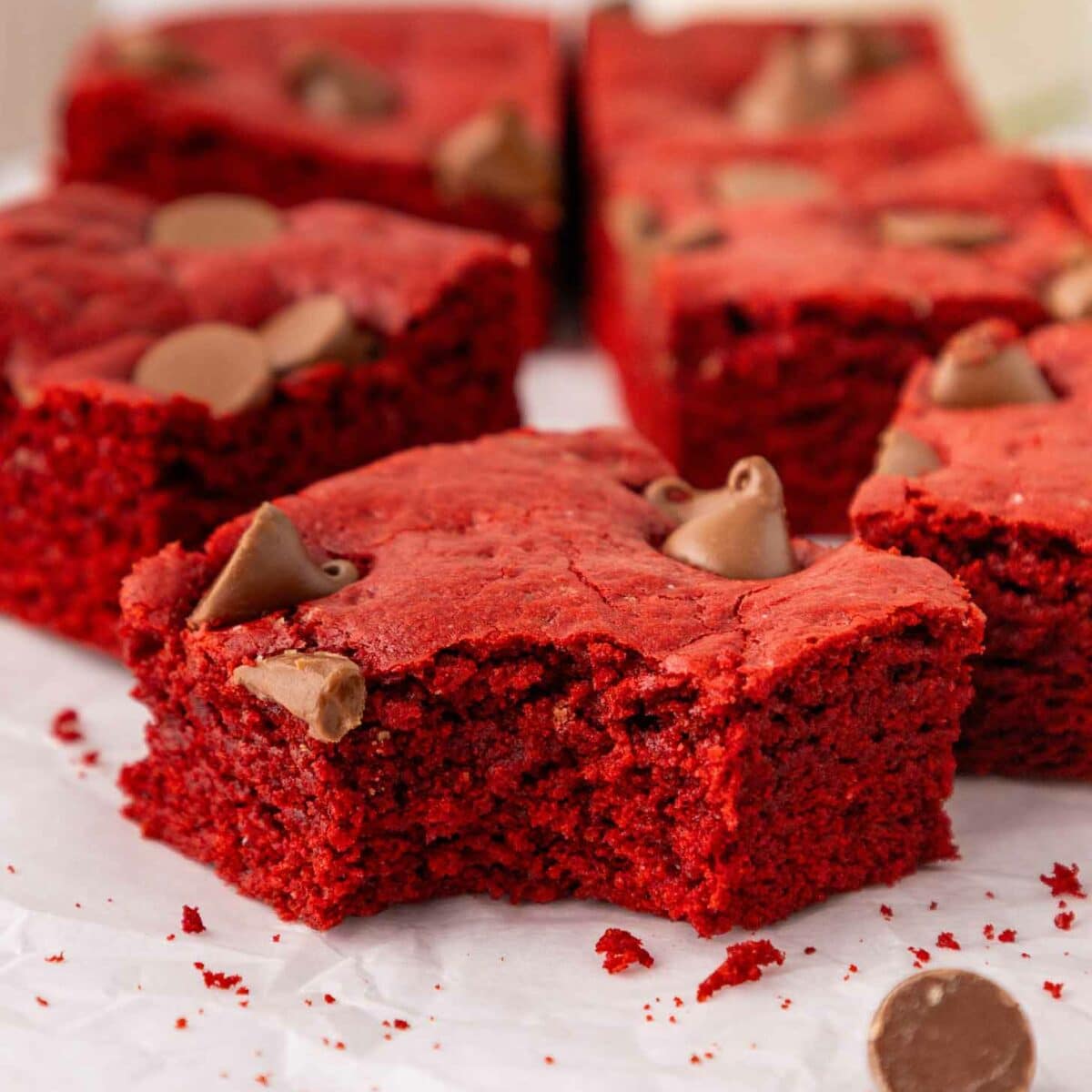 Red velvet brownies topped with chocolate chips are arranged on parchment paper, with one brownie in the foreground showing a large bite taken out of it and scattered crumbs around.