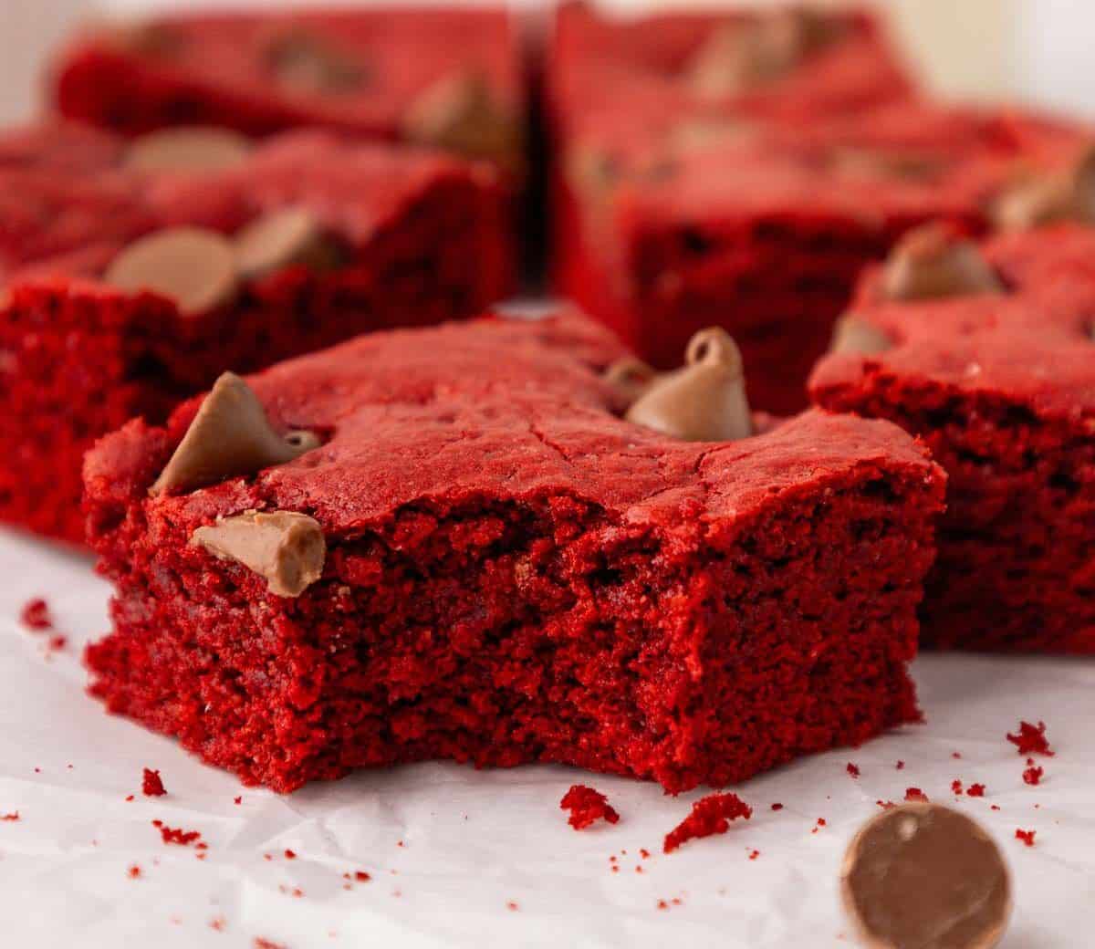 A close-up of a red velvet brownie with a bite taken out, topped with chocolate chips, resting on parchment paper with other brownies and scattered chips in the background.