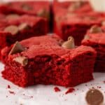 A close-up of a red velvet brownie with a bite taken out, topped with chocolate chips, resting on parchment paper with other brownies and scattered chips in the background.