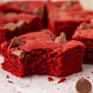 A close-up of a red velvet brownie with a bite taken out, topped with chocolate chips, resting on parchment paper with other brownies and scattered chips in the background.
