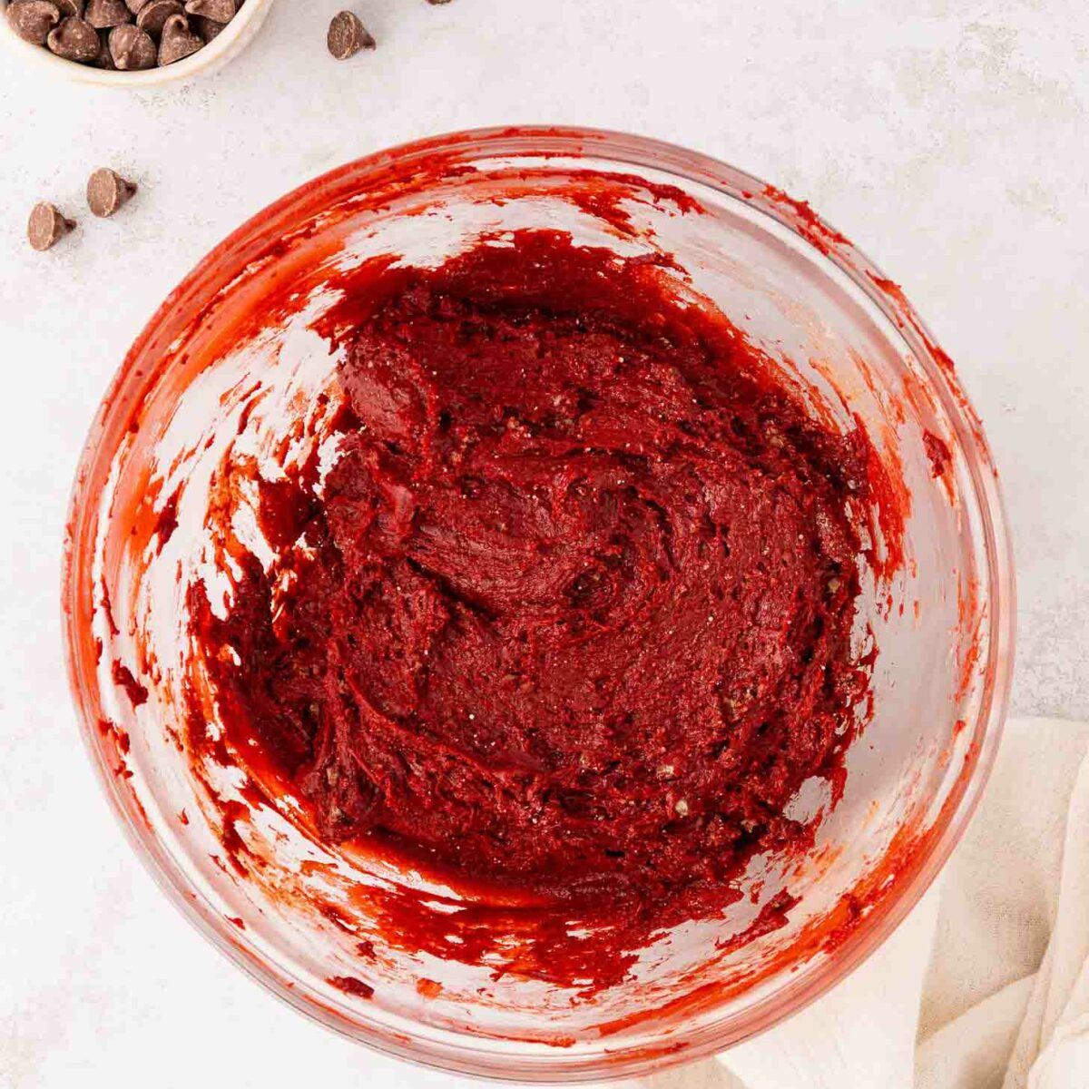A glass bowl filled with red velvet cake mix sits on a light surface, with chocolate chips scattered nearby and a small bowl of chocolate chips in the corner. A cream-colored cloth is partially visible at the bottom right.