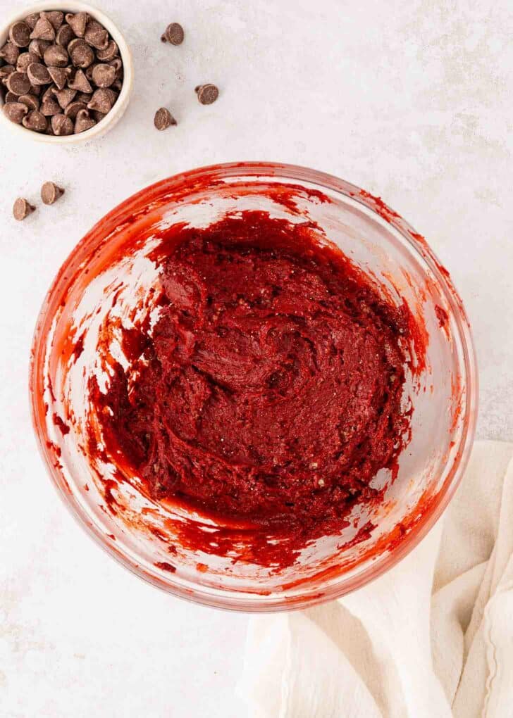 A glass bowl filled with red velvet cake mix sits on a light surface, with chocolate chips scattered nearby and a small bowl of chocolate chips in the corner. A cream-colored cloth is partially visible at the bottom right.