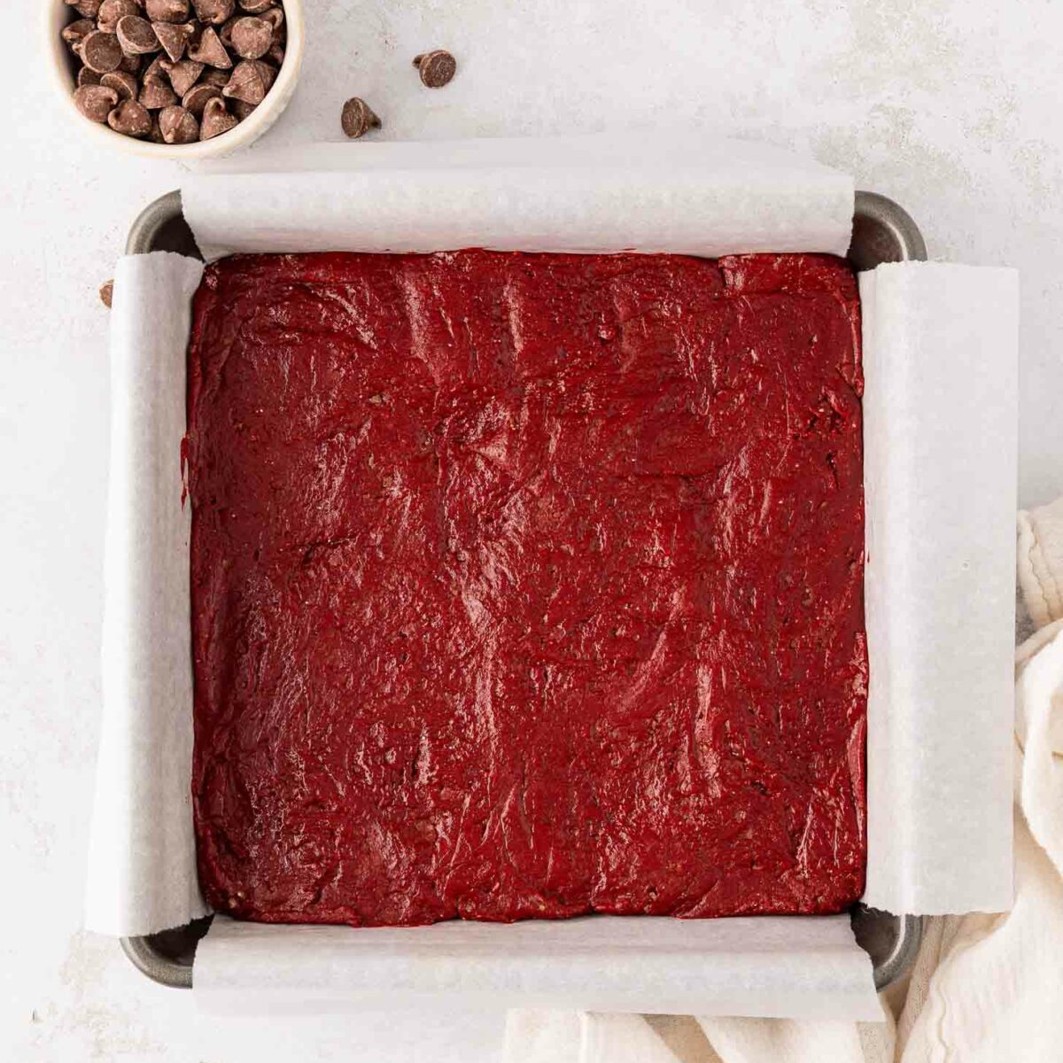 A square baking pan lined with parchment paper is filled with unbaked red velvet batter. A small bowl of chocolate chips sits nearby on a light surface, with a cream-colored cloth partially visible.