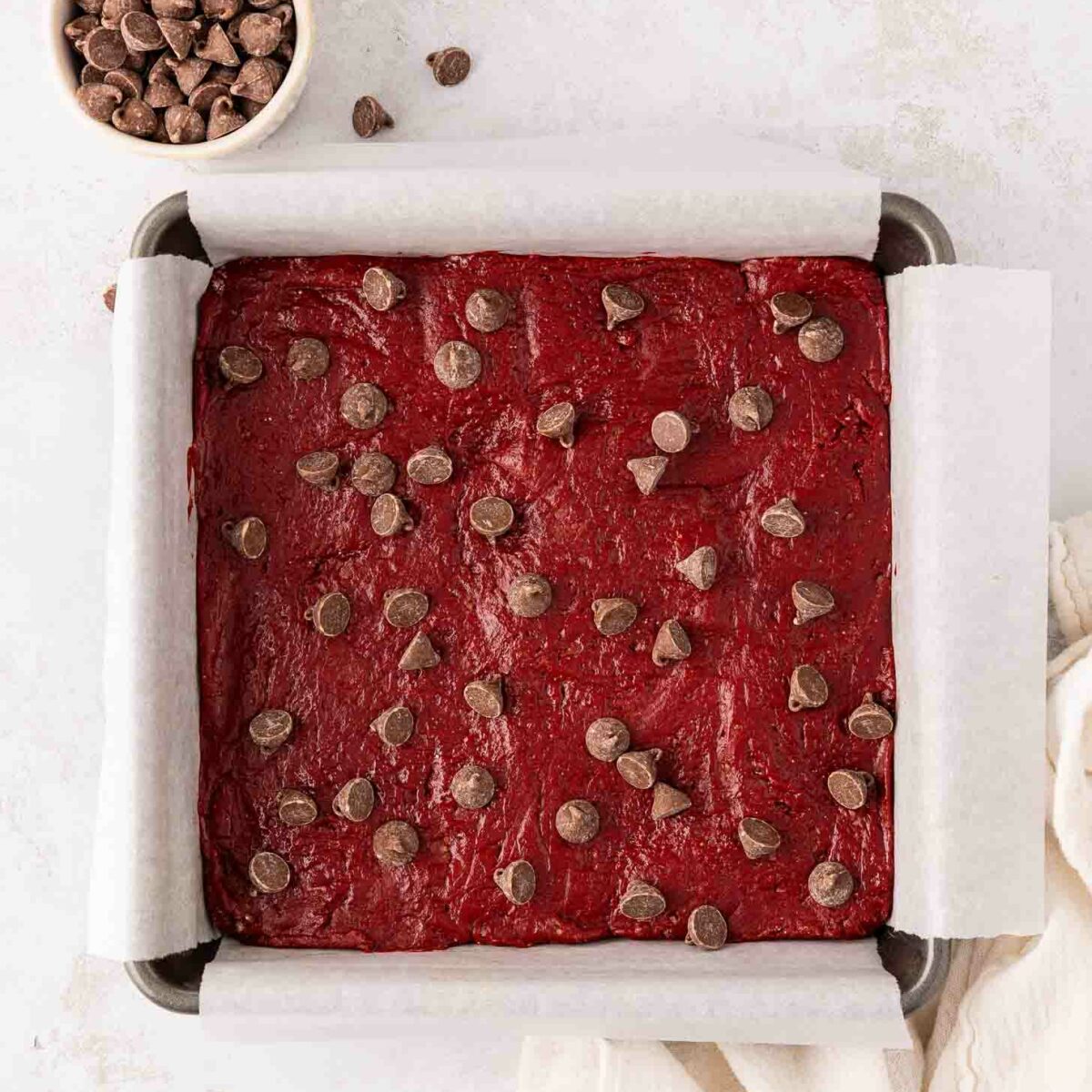 A square baking pan lined with parchment paper is filled with unbaked red velvet brownie batter topped with chocolate chips. A small bowl of chocolate chips sits nearby on a white surface.