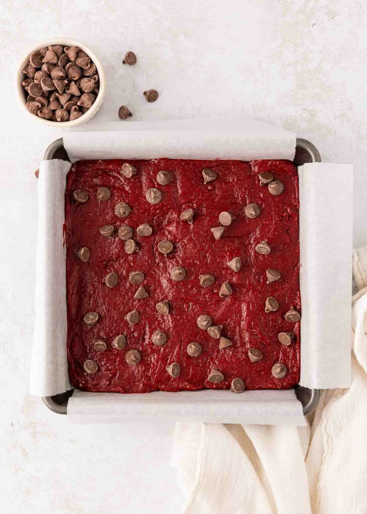 A square baking pan lined with parchment paper is filled with unbaked red velvet brownie batter topped with chocolate chips. A small bowl of chocolate chips sits nearby on a white surface.