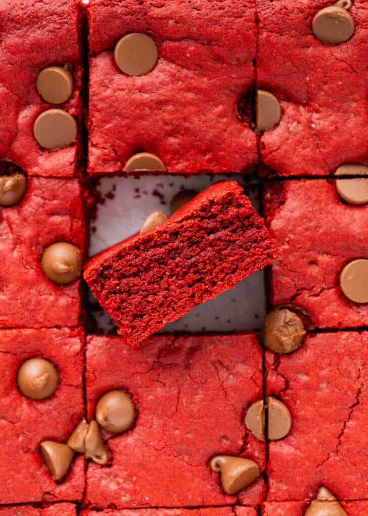 Squares of red velvet brownies topped with milk chocolate chips, with one brownie turned on its side in the center to show its moist, red interior.