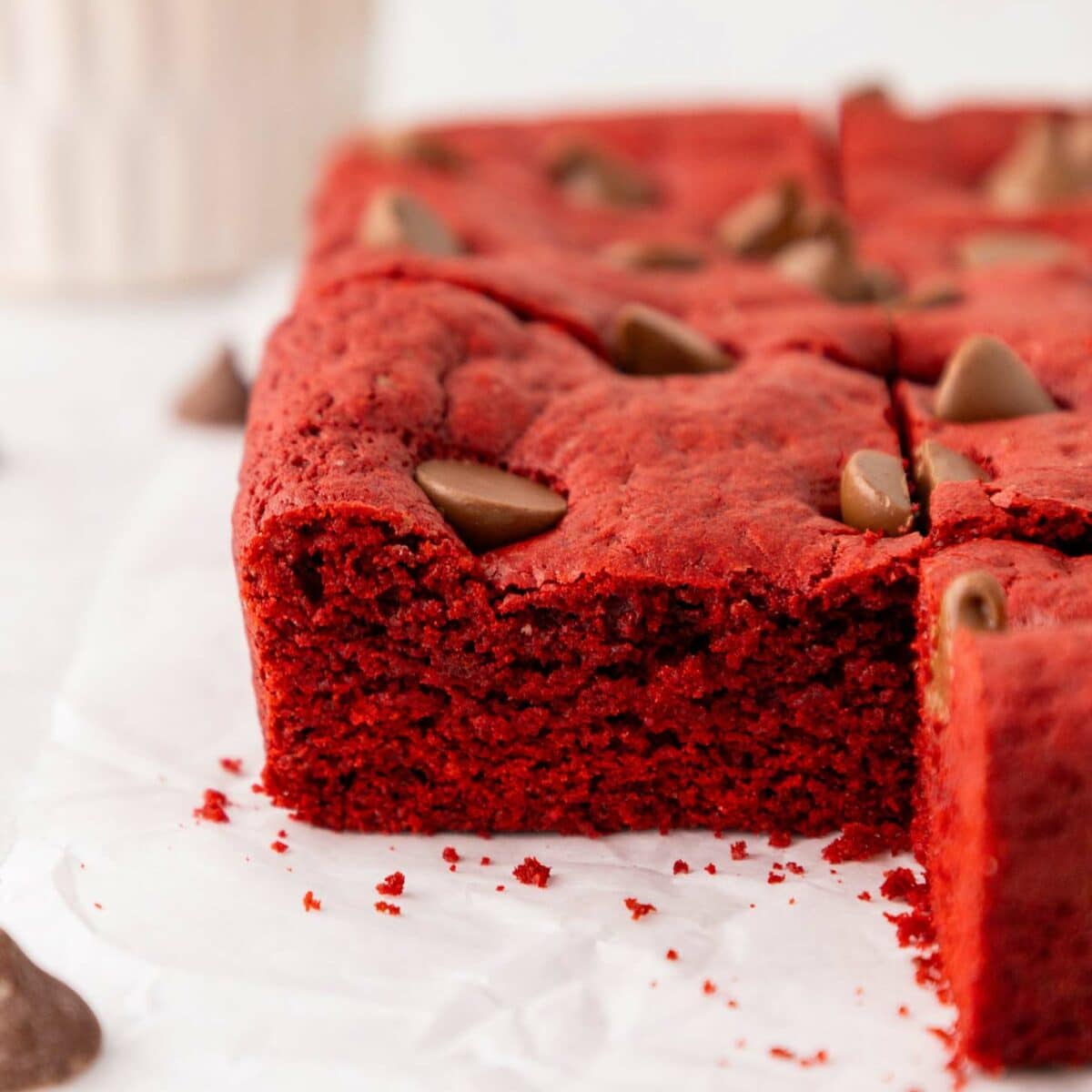 A close-up of thick, red velvet brownies with chocolate chips on top, one piece slightly pulled out, sitting on white parchment paper. A blurred cup and chocolate chips are in the background.
