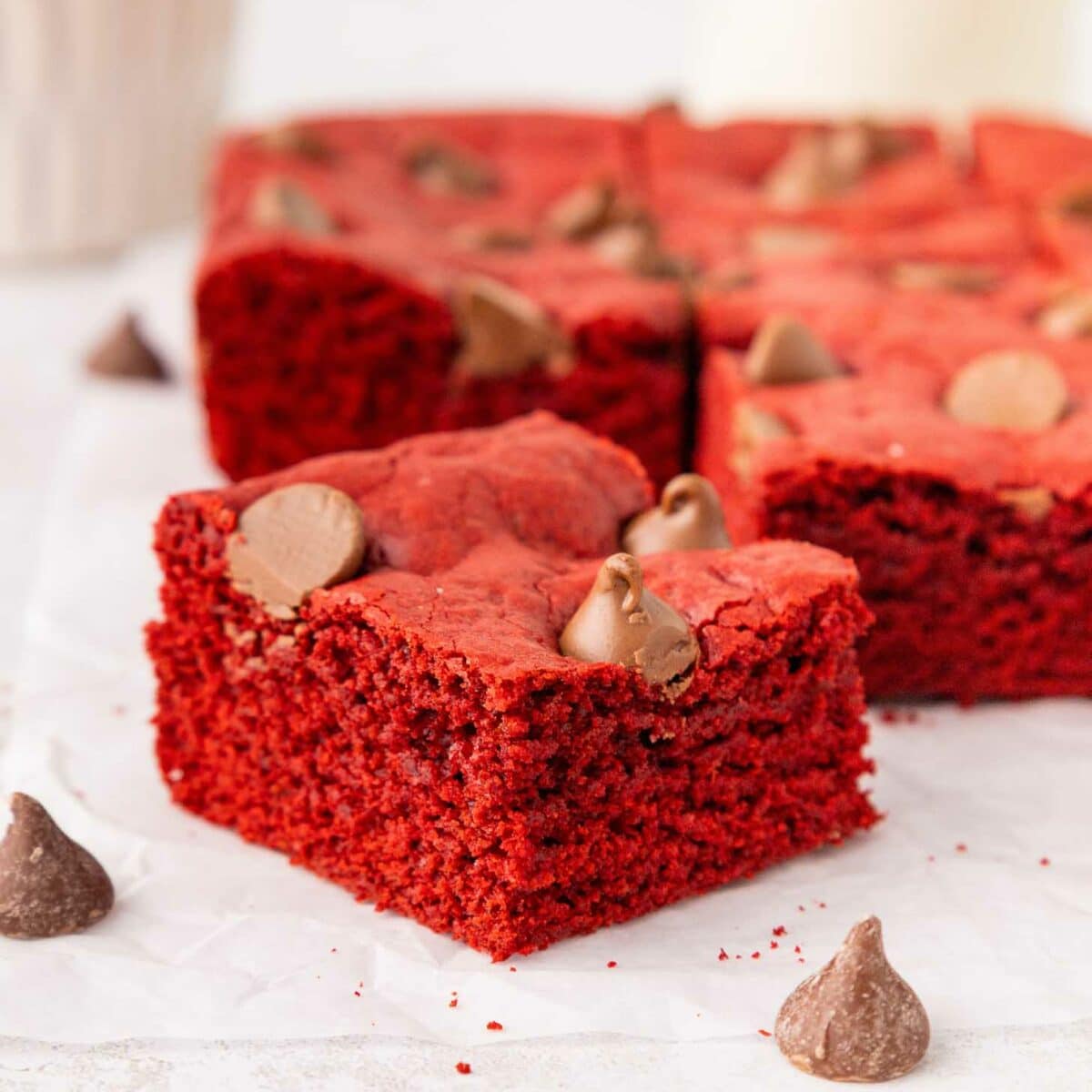 A close-up of square red velvet brownies topped with chocolate chips, arranged on white parchment paper. Chocolate chips are scattered around, and more brownies are blurred in the background.
