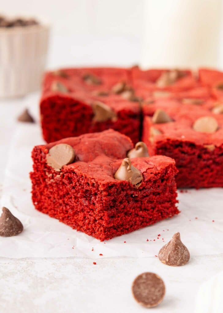 A close-up of square red velvet brownies topped with chocolate chips, arranged on white parchment paper. Chocolate chips are scattered around, and more brownies are blurred in the background.
