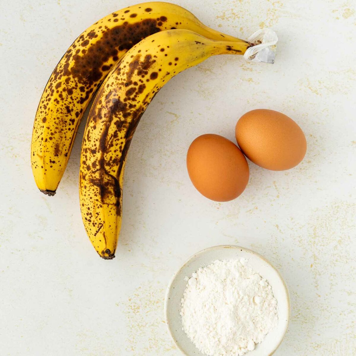 Two ripe bananas with brown spots, two brown eggs, and a small white bowl of flour are arranged on a light, textured surface.