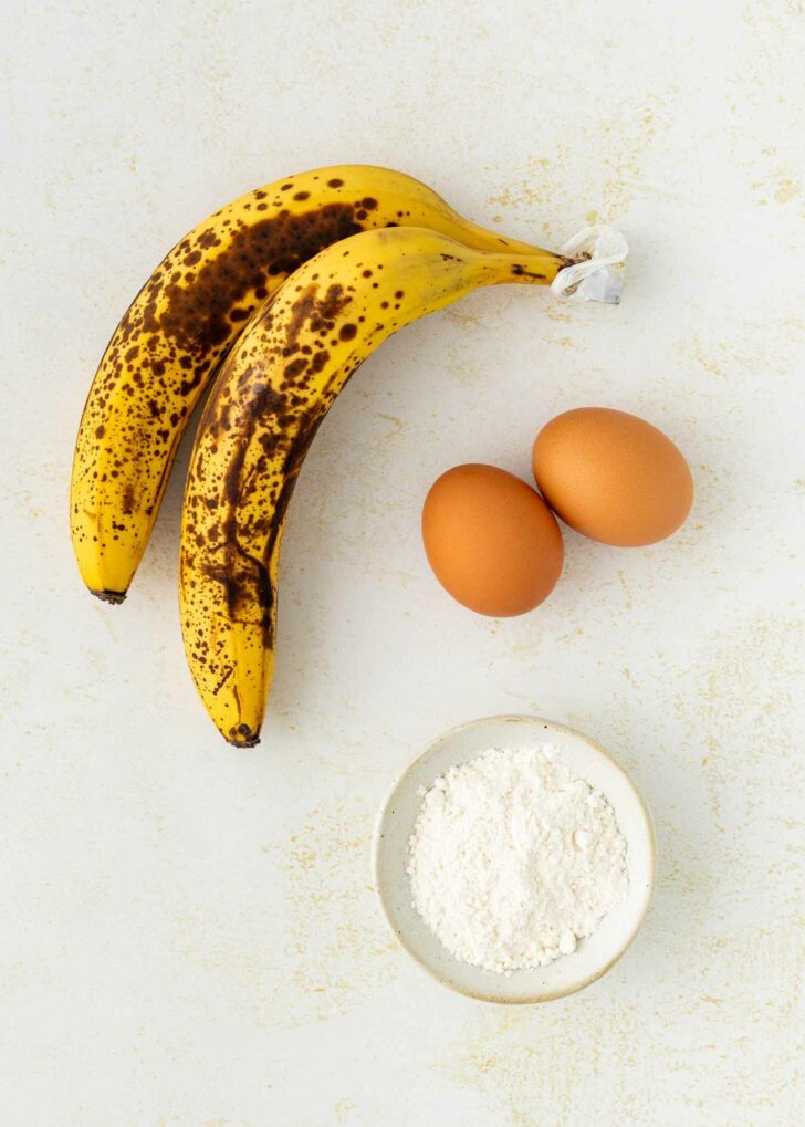 Two ripe bananas with brown spots, two brown eggs, and a small white bowl of flour are arranged on a light, textured surface.